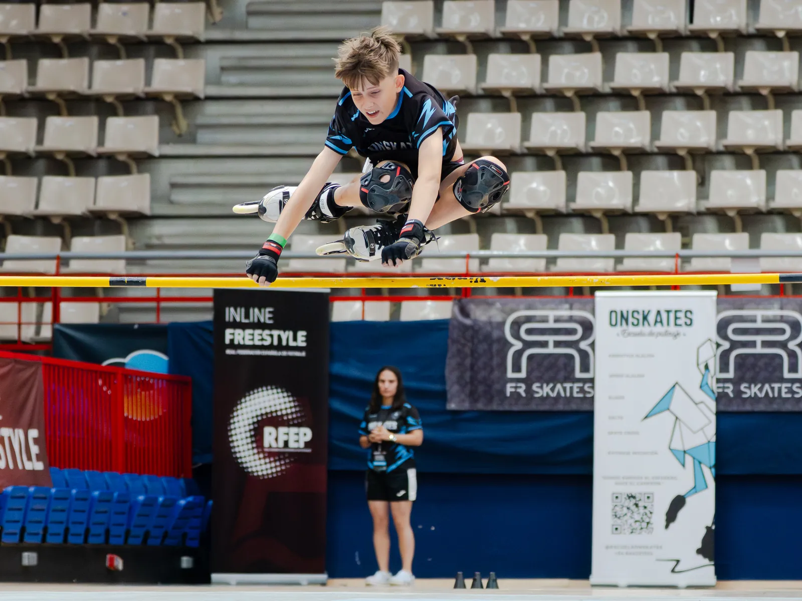 Patinador del Team Onskates en pleno salto de listón en competición, con una entrenadora observando y banners de la RFEP al fondo