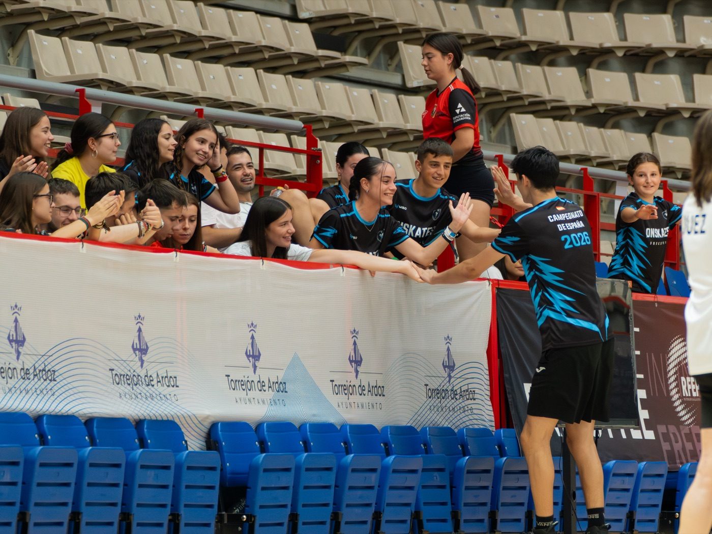 Patinadores del Team Onskates chocando la mano entre el público en el Campeonato de España, banner de Torrejón de Ardoz al fondo