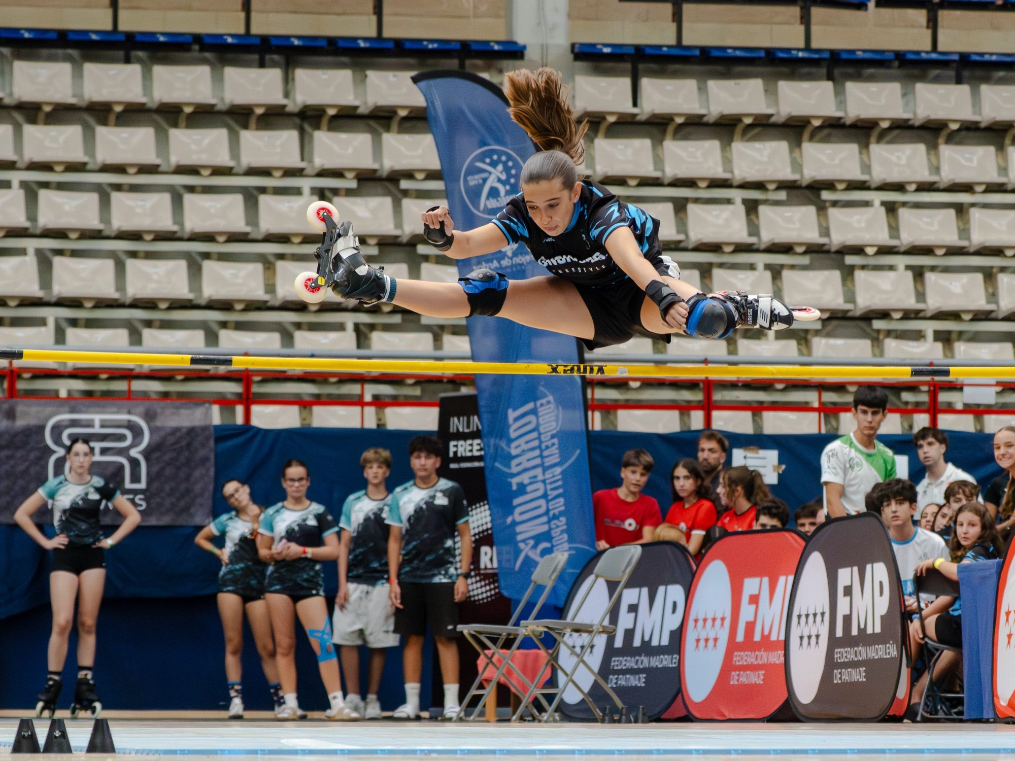 Patinadora del Team Onskates en salto en spagat completo sobre el listón de Free Jump, banners de la Federación Madrileña al fondo